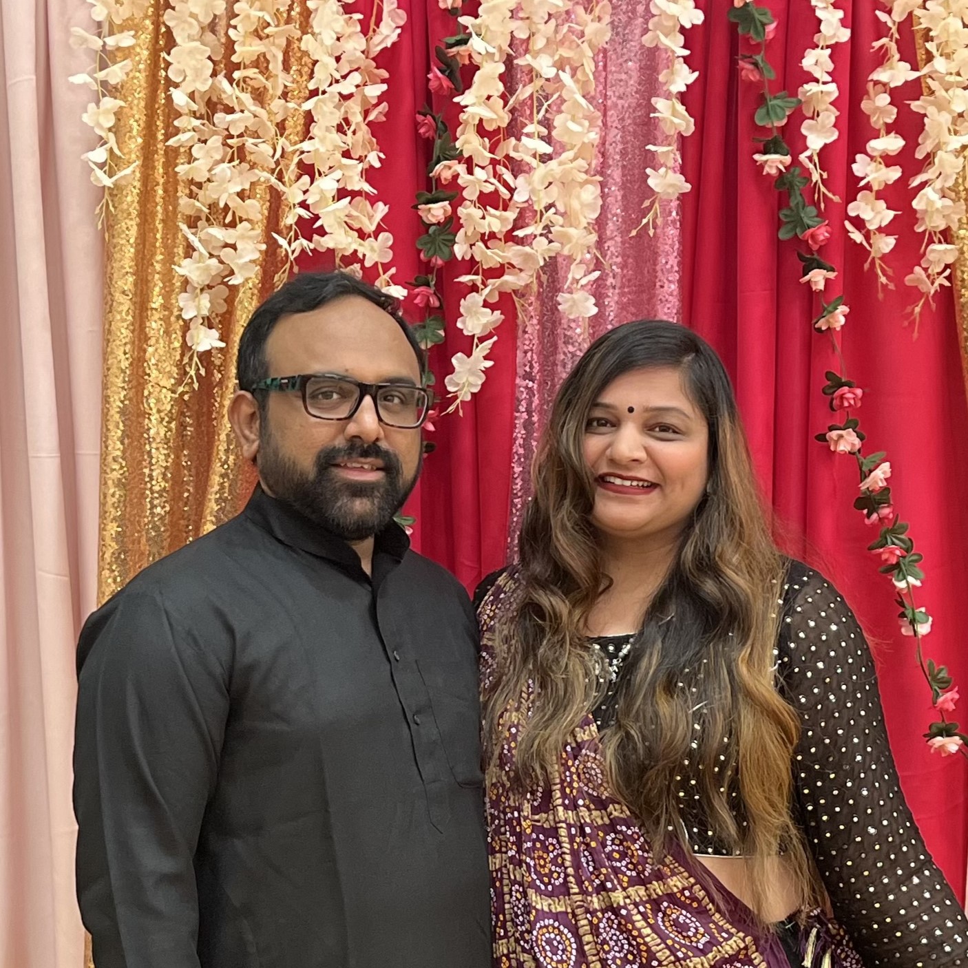 Couple posing joyfully in traditional attire against a vibrant floral backdrop.