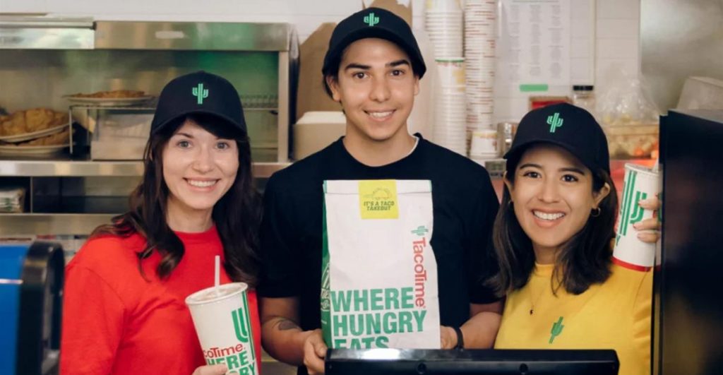 Three smiling TacoTime Canada employees behind the counter, holding TacoTime Canada-branded items / Franchising Mexican Food: A TacoTime Canada Success Story Blog
